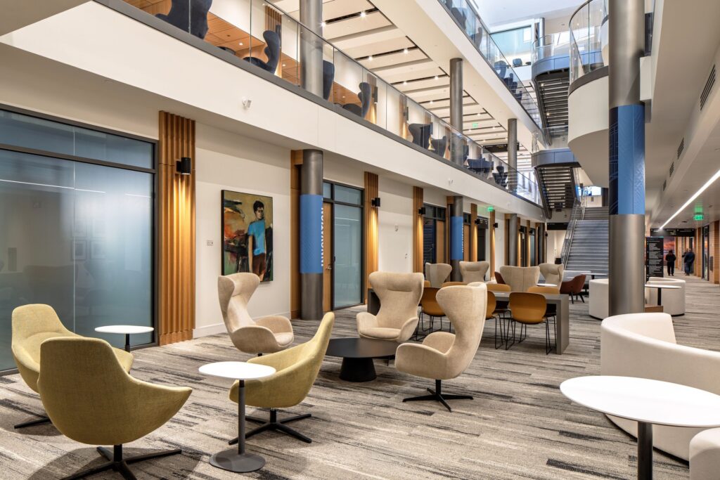 Modern academic building atrium with collaborative seating areas, glass-fronted meeting rooms, and open upper-level walkways.