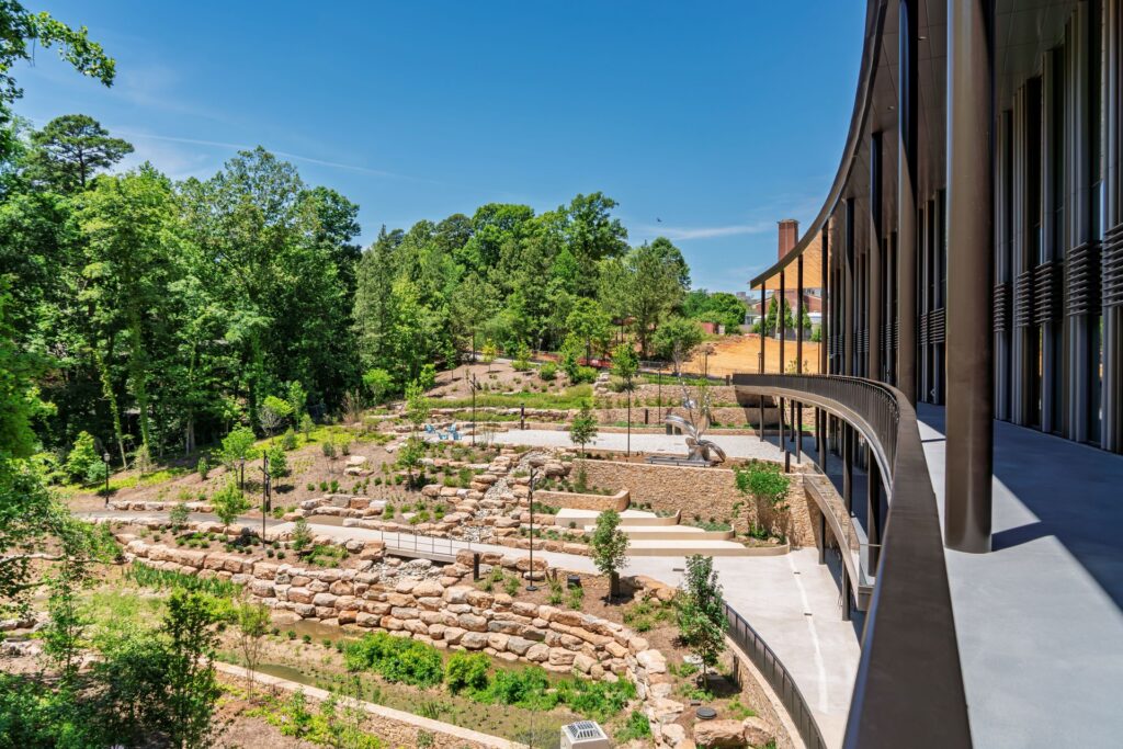 Curved exterior walkway of the UNC Kenan-Flagler Business School addition overlooking a terraced landscape with stone retaining walls, native plantings, and outdoor gathering spaces.