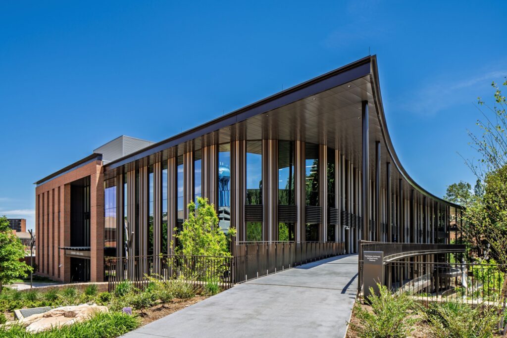 Modern academic building with a curved roofline, floor-to-ceiling glass, vertical sunshades, and an elevated pedestrian walkway leading to the entrance.
