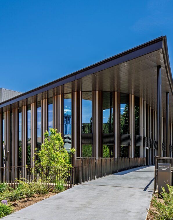 Modern academic building with a curved roofline, floor-to-ceiling glass, vertical sunshades, and an elevated pedestrian walkway leading to the entrance.